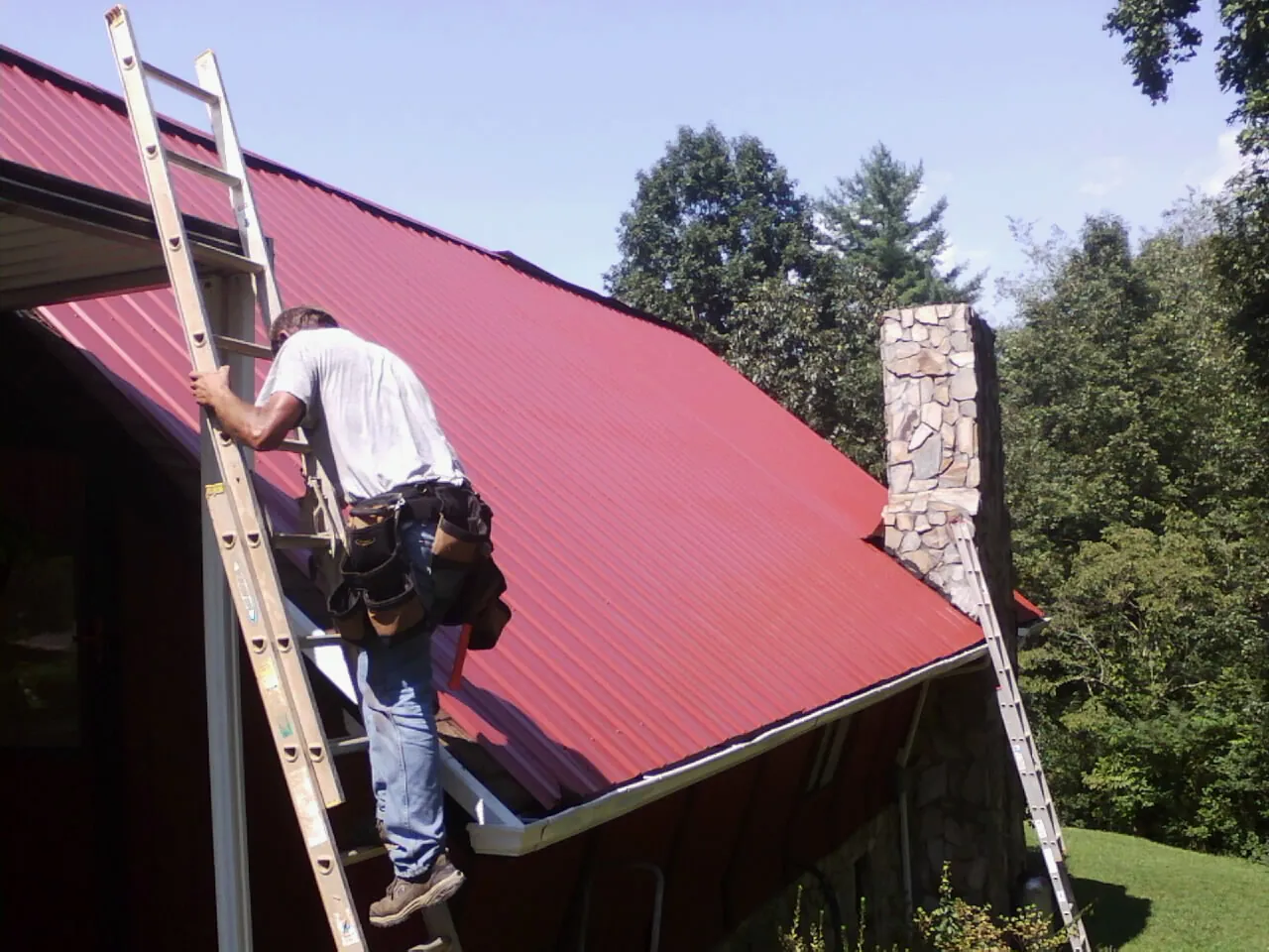 Roofer on a ladder beside a red metal roof and stone chimney — Soffit & Fascia Repair by Roofing Brothers in New Albany